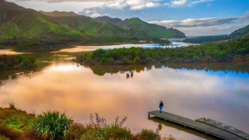Serene Woman Standing on a Wooden Pier Overlooking Golden Lake and Lush Green Mountains 4K Wallpaper