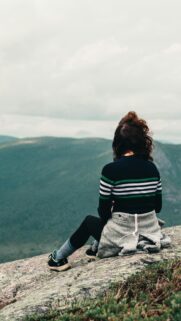 Woman Sitting on Mountain Edge Overlooking Green Valley Wallpaper