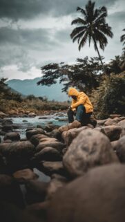 Lone Adventurer in Yellow Hoodie by a Rocky River Wallpaper