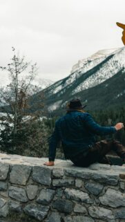 Man in Denim Jacket and Hat Looking at Snowy Mountains Wallpaper