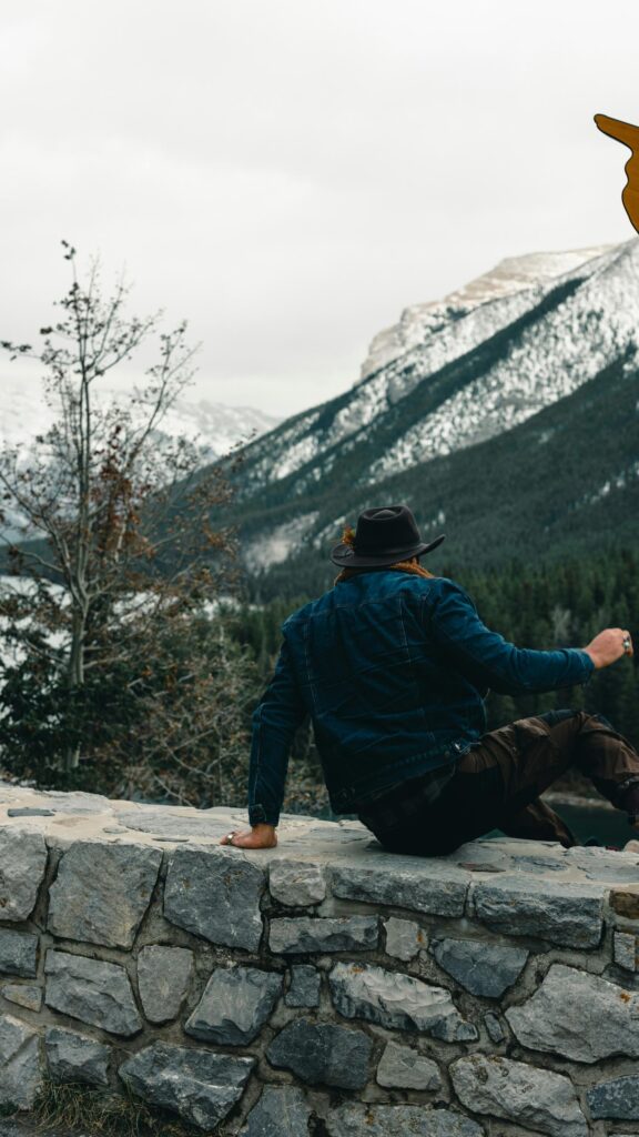Man in Denim Jacket and Hat Looking at Snowy Mountains Wallpaper