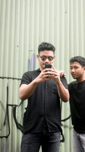 Two Young Men Checking Phone Against Graffiti Metal Wall Wallpaper