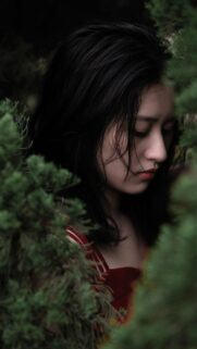 Sad Asian Girl in Red Dress Surrounded by Lush Green Foliage Wallpaper