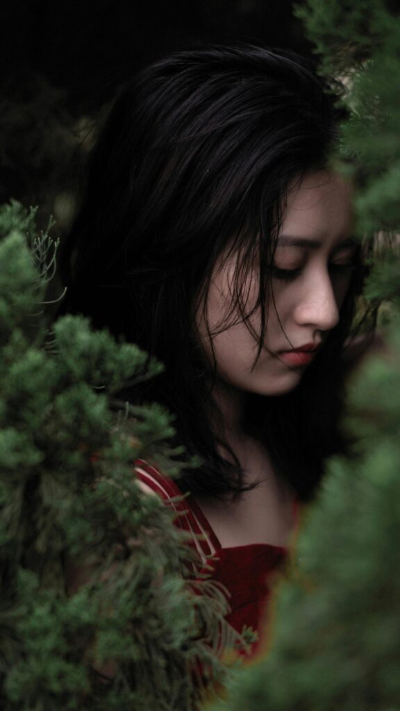 Sad Asian Girl in Red Dress Surrounded by Lush Green Foliage Wallpaper