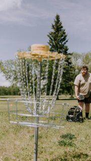 Disc Golf Player Standing by Basket in Sunny Field Phone Wallpaper