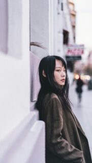 Stunning Asian Woman in Brown Coat Leaning Against a Wall Mobile Wallpaper
