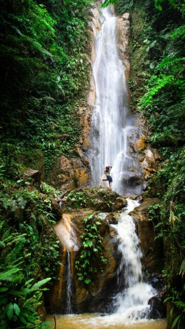 Woman Standing Beneath Majestic Tropical Waterfall Mobile Wallpaper