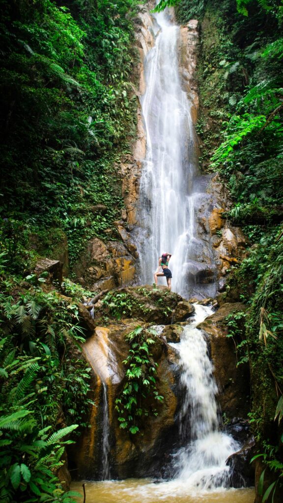 Woman Standing Beneath Majestic Tropical Waterfall Mobile Wallpaper