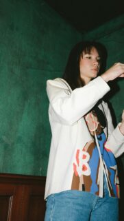 Young Woman in Stylish Graphic Jacket Against Moody Green Wall Wallpaper