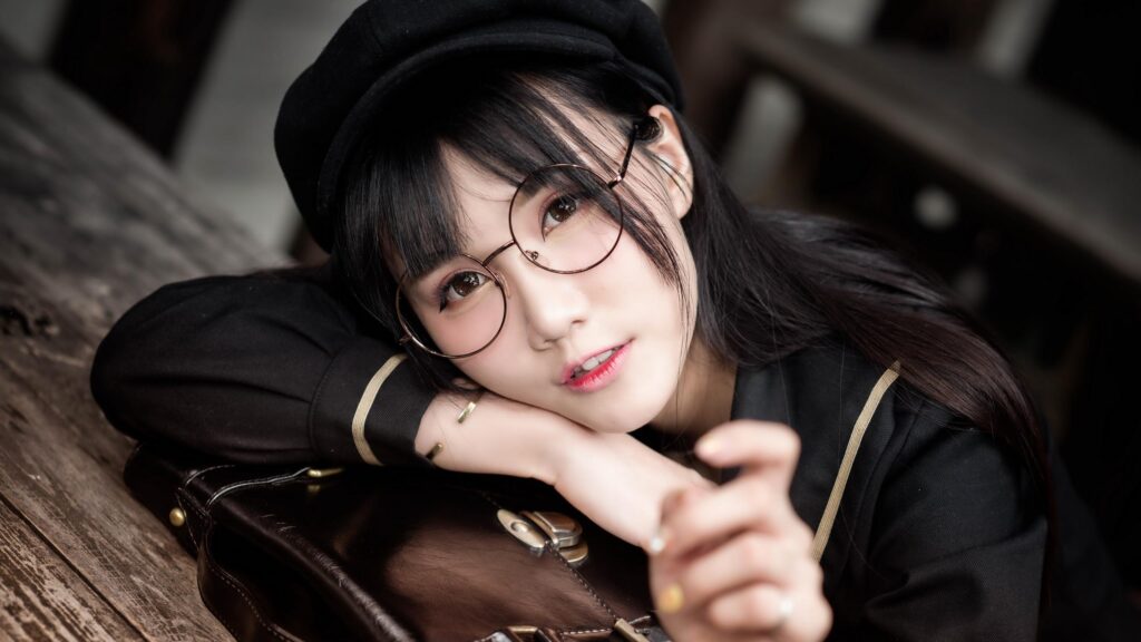 Charming Asian Girl with Glasses and Beret Resting on Vintage Suitcase Desktop Wallpaper