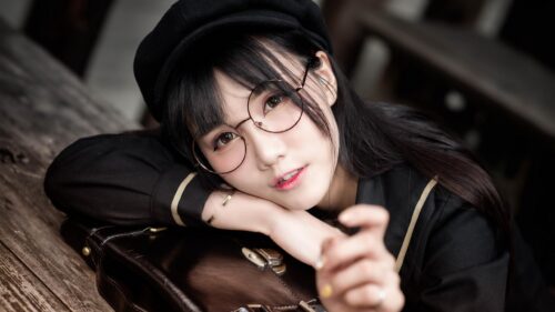 Charming Asian Girl with Glasses and Beret Resting on Vintage Suitcase Desktop Wallpaper