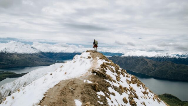 Hiker Standing on Snowy Mountain Peak Overlooking Lake Desktop Wallpaper