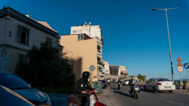 Sunlit Coastal Street with Motorcyclists in Greece Desktop Wallpaper