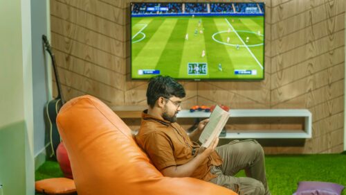 Man Reading Book on Orange Beanbag Sofa During Soccer Game 4K Wallpaper