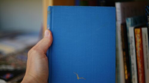 A Person Holding a Vintage Blue Hardcover Book on a Bookshelf Desktop Wallpaper