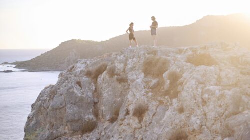 Two Hikers Standing on a Rocky Cliff Overlooking the Sea 4K Wallpaper