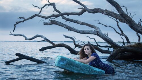 Ethereal Woman Resting on Floating Foam in Serene Ocean Water with Driftwood Backdrop 4K Wallpaper